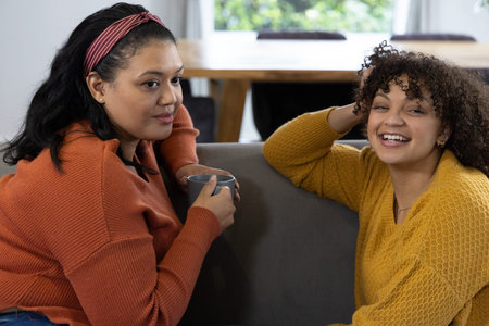 Diverse female friends sitting on grey sofa at home holding grey mug, wearing sweaters. Duo, women, cozy, warm, autumn, knit, headbandの写真素材