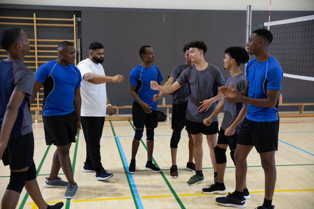 Male teen volleyball team and coach gathering in school gym near net, wearing jerseys, knee pads....