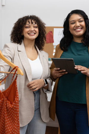 Diverse female team presenting polka-dot dress while consulting tablet and corkboard at boutique....