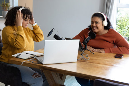 African American and Indian women sitting at table podcasting with laptop mics headsets in knits. Cohosts, microphones, headphones, conversation, casual, home, studioの写真素材