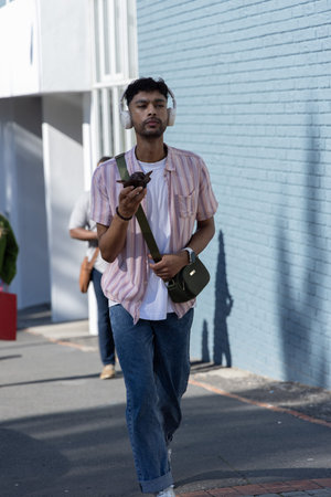 Asian man walking sidewalk by blue brick wall wearing white headphones with bag holding phone....