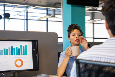 Diverse team African American woman in blue dotted top holding mug talking dashboard charts at desk....