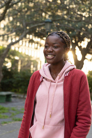 Teenage female smiling, standing on path in park wearing pink hoodie and red jacket, beaded braids. Plaza, lamppost, bench, trees, sunlight, goldenhour, backlightの写真素材