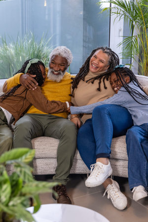 African American family cuddling on sofa at home, senior hugging two child girls by coffee table....