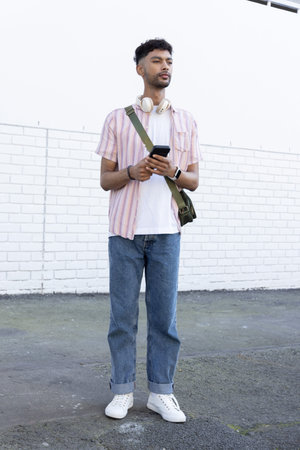 Male standing against white brick wall on paved alley holding smartphone and wearing headphones....