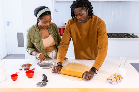 African American couple making dough in kitchen using rolling pin, pie tin, woman wearing headband. Baking, measuring, home, bright, cozy, collaboration, culinaryの写真素材