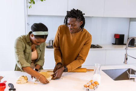 African American couple in casual wear prepping dough with rolling pin and eggs at kitchen island....