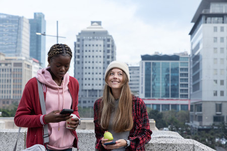Diverse female friends standing at stone railing on city terrace, holding coffee, phones. Urban, outdoor, smartphones, casual, duo, skyline, beanieの写真素材
