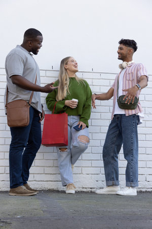 Three friends chatting while standing in front of white painted brick wall holding red shopping bag. People, group, conversation, outdoor, street, sidewalk, urbanの写真素材