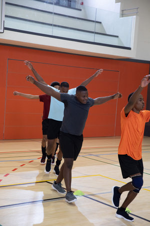 African American male athletes wearing shirts and shorts doing agility drill at gym with green cone....