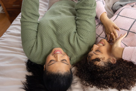 Diverse female friends lying on bed facing each other, wearing green knit and pink check sweaters. Women, mattress, bedroom, cozy, intimate, friendship, comfortの写真素材