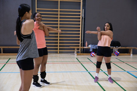 Diverse teenage female volleyball players stretching on gym court near wall bars and volleyballs....