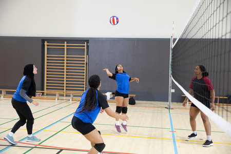 Four female volleyball players jumping, hitting volleyball at net on gym court in blue jerseys....