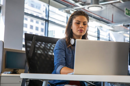 Woman sitting in black mesh chair at white office desk using silver laptop wearing white headphones....
