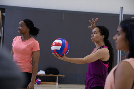 Diverse female teens serving at gym, server in purple tank holding red-white-blue ball, copy space. Volleyball, teammates, gymnasium, hardwood, court, practice, dynamicの写真素材