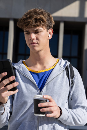 Teen male wearing gray hoodie and blue shirt standing at campus holding smartphone and coffee....