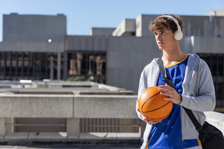 Teenage male standing on rooftop holding orange basketball, wearing blue jersey, copy space....