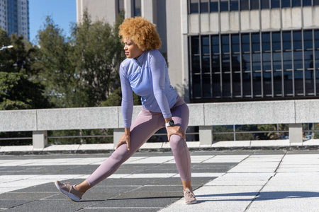 African American woman doing wide side lunge on rooftop plaza, wearing lilac top. Fitness, outdoor,...
