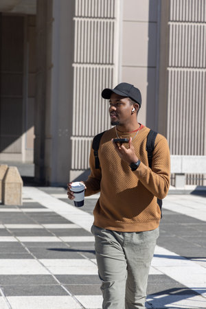 African American man walking across urban plaza while speaking into smartphone and holding coffee....