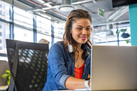 Woman working on laptop at desk in modern open-plan office, wearing denim shirt, headphones....