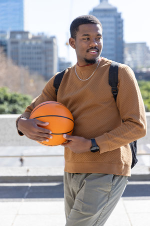 African American man holding basketball, standing on plaza with black backpack, in brown sweatshirt. Athlete, urban, outdoor, candid, streetstyle, casual, cityscapeの写真素材