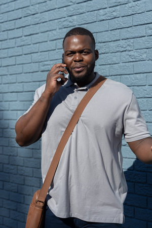 African American man 30s standing, talking on phone against blue brick wall with gray polo, satchel....