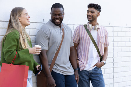 Diverse friends leaning on painted white brick wall, laughing, holding coffee cup, red bag. Trio,...