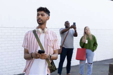 Diverse friends standing on sidewalk holding smartphone, headphones, coffee. Urban, street, group,...
