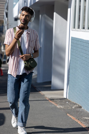 Asian man walking on sunlit sidewalk inspecting chocolate muffin, wearing headphones. Pedestrian, urban, street, casual, earbuds, satchelの写真素材