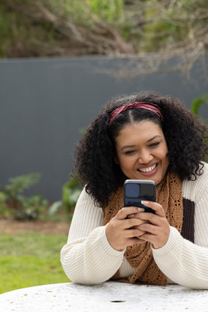 African American woman sitting at round white table in garden, wearing cream sweater, holding phone....