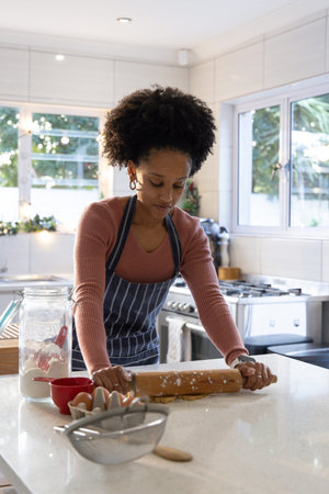 African American woman in striped apron rolling dough on quartz counter with rolling pin, flour jar....