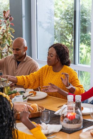 Diverse family sharing meal at table by windows African American woman in yellow top reaching rolls....