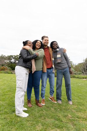 Diverse friends hugging and laughing while standing on park lawn wearing jackets and scarf. Group,...