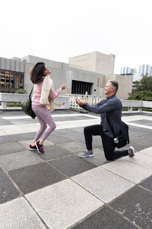 Couple on rooftop terrace with man kneeling, presenting open box woman in pink jacket raising hands....