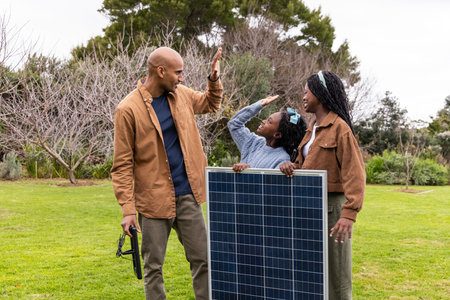 African American family wearing brown coats by solar panel, man highfiving checking tool on lawn....