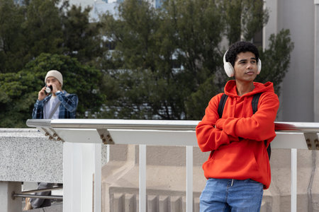 Two teen males on balcony, wearing red hoodie and headphones, other sipping coffee, copy space. Urban, street, concrete, railing, backpack, casualの写真素材