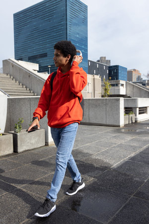 African American teenage male walking across wet plaza in red hoodie and backpack adjusting headset....
