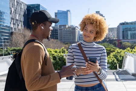 Diverse friends chatting on terrace, man with backpack holding mug, woman holding smartphone. Urban,...
