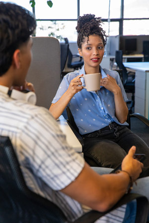 Diverse coworkers chatting at adjacent desks in open-plan office, holding beige mug and smartphone....