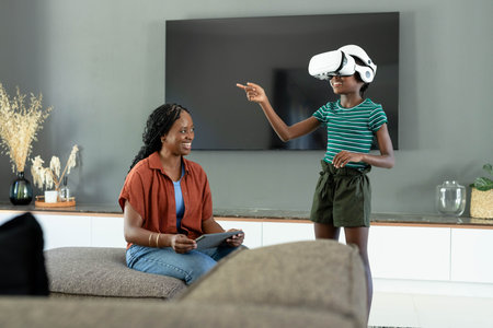 African American mom holding tablet, daughter wearing white VR headset pointing at home, copy space. Family, woman, child, goggles, screen, livingroom, modernの写真素材