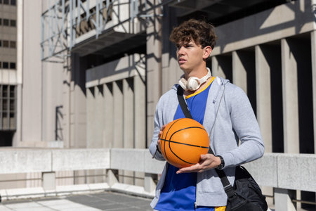 Teenage male standing holding basketball in blue jersey, white headphones, black duffel on rooftop. Urban, architectural, athletic, outdoor, sunlit, focus, determinationの写真素材