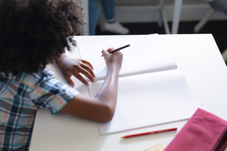 African american elementary schoolboy writing on book at desk in classroom. unaltered, education,...