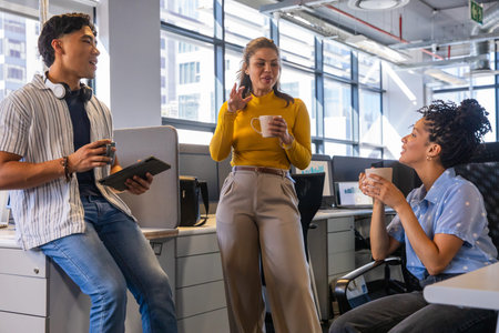 Diverse coworkers chatting, holding coffee mugs and tablet near large windows in modern office....
