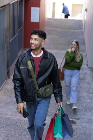 Indian man walking up alley wearing leather jacket carrying colorful bags and using smartphone. Person, urban, street, passage, ramp, phone, casualの写真素材