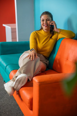 Woman leaning on teal sofa, holding phone to ear, wearing mustard top, smartwatch in office lounge....