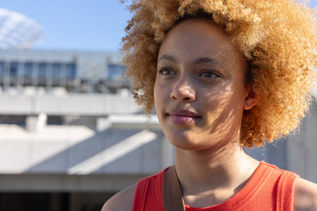 African American woman in her 20s standing by a garage, wearing a red top and carrying a bag strap. Portrait, urban, outdoor, sunlight, architecture, concrete, crimson.の写真素材