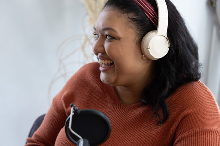 Asian woman smiling, speaking into mic at home studio wearing white headphones and maroon headband. Podcaster, microphone, boomarm, popfilter, creative, content, productionの写真素材