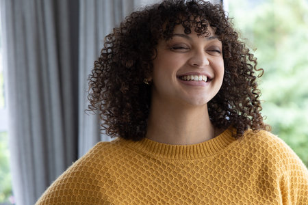 African American woman smiling, wearing mustard-yellow knit sweater, tilting head by window....