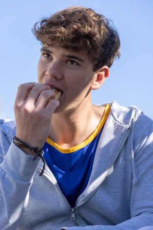 Teenage male biting sandwich wearing blue jersey yellow trim and gray hoodie with bracelets outside....