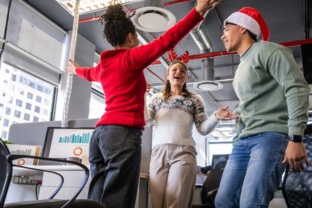 Diverse coworkers celebrating near monitors showing charts in office wearing Santa hat and antlers. Colleagues, festive, celebration, daylight, openplan, cityscape, modernの写真素材